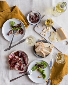 Verbena de San Juan: Jamón Ibérico de Bellota, Foie,Chorizo dulce, fuet y Queso de Cabra, presentados en mesa sobre platos y mantelería blancos. Copas d evino y cubiertos