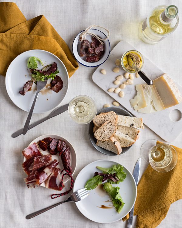 Verbena de San Juan: Jamón Ibérico de Bellota, Foie,Chorizo dulce, fuet y Queso de Cabra, presentados en mesa sobre platos y mantelería blancos. Copas d evino y cubiertos